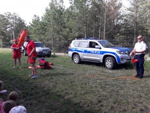 Policjant i ratownicy wodni na tle radiowozu przemawiają do grupy dzieci