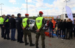 mężczyźni w mundurach policji i żandarmerii wojskowej oraz grupa młodzieży przyglądają się przeprowadzaniu pierwszej pomocy na fantomie