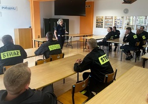 mężczyźni w mundurach straży pożarnej siedzą w ławkach i patrzą na stojącą przed nimi policjantkę prowadzącą szkolenie