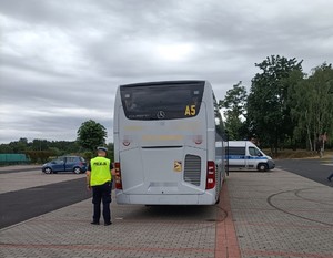 Policjant w kamizelce odblaskowej z napisem POLICJA ogląda z zewnątrz kontrolowany siwy autokar