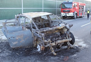 na pierwszym planie widać stojące na jezdni spalone auto, w tle policjant