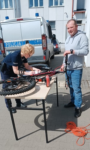 Policjantka znakuje rower. Przed  nią stoi właściciel jednośladu. Rower położony jest na stole.