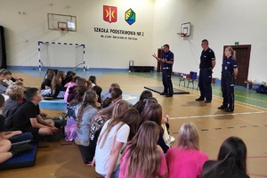 Policjanci prowadzący spotkanie z młodzieżą na sali gimnastycznej szkoły podstawowej, na zdjęciach dzieci z hulajnogami i kaskami ochronnymi