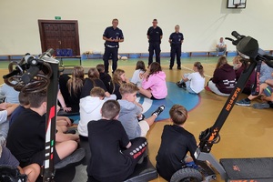 Policjanci prowadzący spotkanie z młodzieżą na sali gimnastycznej szkoły podstawowej, na zdjęciach dzieci z hulajnogami i kaskami ochronnymi
