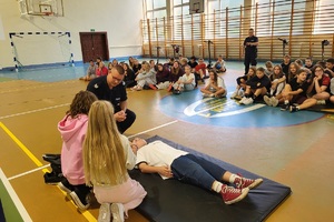 Policjanci prowadzący spotkanie z młodzieżą na sali gimnastycznej szkoły podstawowej, na zdjęciach dzieci z hulajnogami i kaskami ochronnymi