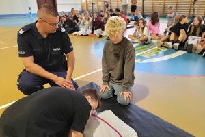 Policjanci prowadzący spotkanie z młodzieżą na sali gimnastycznej szkoły podstawowej, na zdjęciach dzieci z hulajnogami i kaskami ochronnymi
