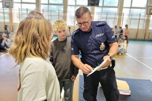 Policjanci prowadzący spotkanie z młodzieżą na sali gimnastycznej szkoły podstawowej, na zdjęciach dzieci z hulajnogami i kaskami ochronnymi