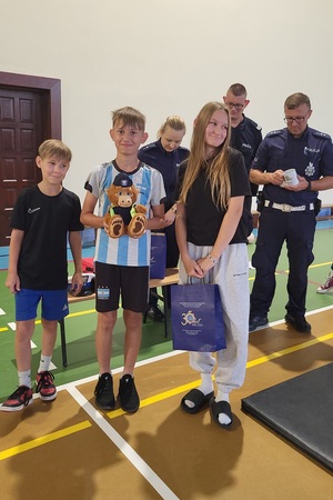 Policjanci prowadzący spotkanie z młodzieżą na sali gimnastycznej szkoły podstawowej, na zdjęciach dzieci z hulajnogami i kaskami ochronnymi