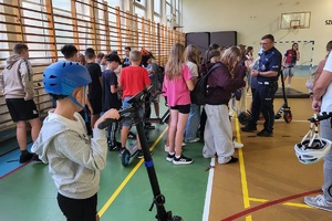 Policjanci prowadzący spotkanie z młodzieżą na sali gimnastycznej szkoły podstawowej, na zdjęciach dzieci z hulajnogami i kaskami ochronnymi