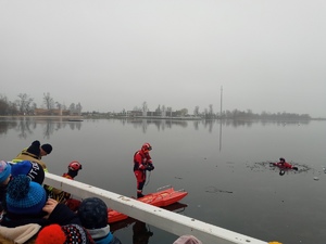 Pokaz ratownictwa wodnego na lodzie. Na dole fotografii widoczne głowy dzieci, które są zwrócone w kierunku tafli lodowej. Lodzie znajduje się 3 strażaków, którzy prowadzą pokaz, jak udzielić pomocy tonącemu. Mają ze sobą specjalistyczna deskę ratunkową.