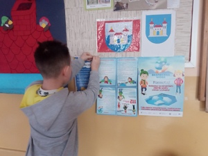 Uczeń przypina na gazetce informacje promujące bezpieczne zachowania podczas ferii zimowych. Na ścianie zawieszone są rożne kolorowe prace wykonane przez uczniów. Chłopiec stoi tyłem do autora zdjęcia.
