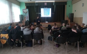 Zdjęcie grupowe. Sala w rogowskim domu kultury. Na pierwszym planie tyłem do autora zdjęcia siedzą dzieci na krzesłach. Przed nimi stoi policjantka sierż. szt. Katarzyna  Leszczyńska - Krawczyk. z jej prawej strony wyświetlana na rzutniku jest prezentacja.